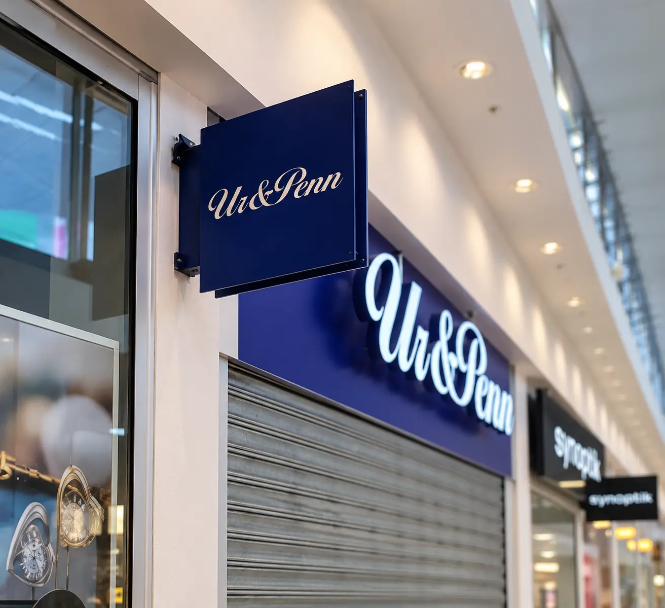 Blå dubbelsidig flaggskylt för Ur & Penn monterad vinkelrätt från butiksfasaden